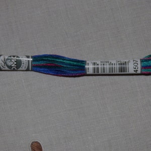 Puede incluir: Hilo de bordar DMC en tonos de azul verdoso, azul, rosa y morado. El hilo está envuelto alrededor de un carrete de cartón blanco con un código de barras en blanco y negro y el número 4507.