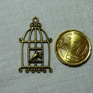 Puede incluir: Un charm de jaula de pájaros de color bronce con un pájaro dentro, junto a una moneda de 20 céntimos. El charm tiene una parte superior arqueada y remolinos decorativos. La moneda tiene aproximadamente 2,4 cm de diámetro. El fondo es de tela gris claro.