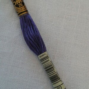 Könnte beinhalten: Ein Knäuel DMC Stickgarn in Farbe 32, einem tiefen Lilaton. Das Garn ist um eine Pappspule mit einem goldenen Etikett gewickelt, auf dem "DMC" und "25" steht, und hat einen Barcode und die Nummer "32" aufgedruckt.