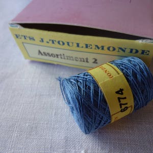 Puede incluir: Una bobina de hilo de bordar azul con una etiqueta amarilla que dice "6774" y "LONDRES". La etiqueta est&aacute; unida a la bobina con un hilo blanco. La bobina est&aacute; sobre una superficie de tela blanca. La bobina es de la empresa "ETS J. TOULEMONDE" y la etiqueta dice "Assortiment 2".