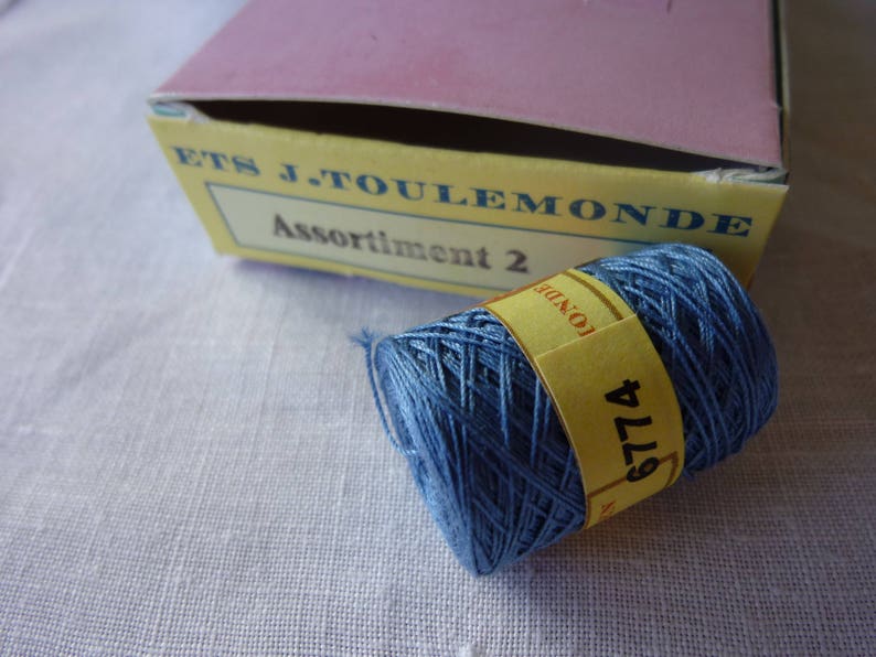 Puede incluir: Una bobina de hilo de bordar azul con una etiqueta amarilla que dice "6774" y "LONDRES". La etiqueta est&aacute; unida a la bobina con un hilo blanco. La bobina est&aacute; sobre una superficie de tela blanca. La bobina es de la empresa "ETS J. TOULEMONDE" y la etiqueta dice "Assortiment 2".