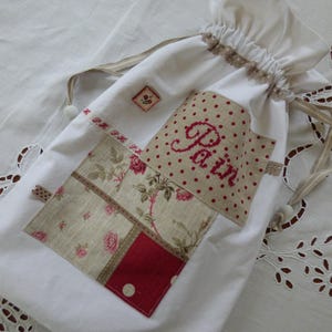 Puede incluir: Bolsa blanca con cordón y patchwork floral con la palabra "Pain" bordada en rojo. La bolsa tiene un ribete decorativo y un pequeño cuadrado bordado. La bolsa está sobre una superficie blanca.