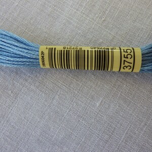 Puede incluir: Un ovillo de hilo de bordar azul claro con una etiqueta amarilla que dice "3755" y un código de barras. La etiqueta también tiene el número "3906042P" impreso.