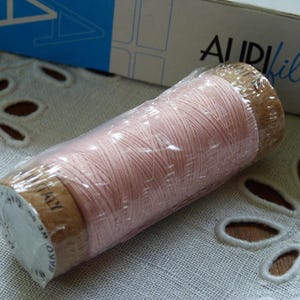 May include: A spool of light pink thread, wrapped in clear plastic, sits on a white embroidered surface. The thread is labeled "Made in Italy". A partially visible box in the background reads "Aurifil".