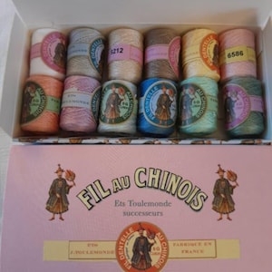 Può includere: Una scatola rosa di rocchetti di filo Fil Au Chinois. La scatola presenta illustrazioni di un uomo in abiti tradizionali. Le bobine sono in vari colori, tra cui bianco, rosa, beige, marrone, giallo e verde. Il testo "Fil Au Chinois" è visibile.