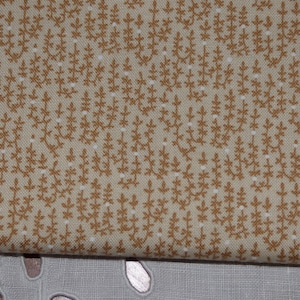 Può includere: Primo piano di un tessuto beige con un motivo ripetuto di piccoli rametti floreali marroni. Il tessuto ha un aspetto leggermente strutturato e una palette di colori morbidi e neutri. Il design ricorda i tessuti vintage o in stile rustico.