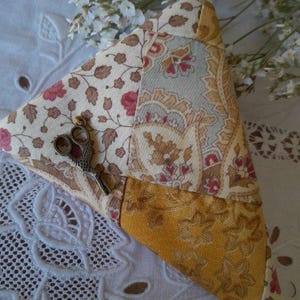 Peut inclure: Un coussin triangulaire en patchwork avec des motifs crème, jaune et floraux. Une petite paire de ciseaux argentés repose sur le coussin. Le coussin est posé sur une surface blanche brodée.