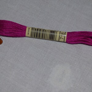 Op de afbeelding: Een spoel levendig magenta borduurgaren, gemarkeerd met nummer 718. Het garen is strak opgerold en heeft een lichte glans. Het etiket bevat een barcode en andere gedrukte informatie.