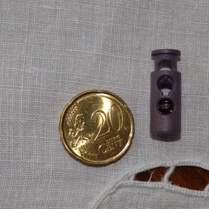 May include: A purple plastic cord lock with a metal slider, next to a 20 euro cent coin.