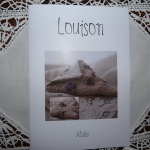 Puede incluir: Una tarjeta blanca con la palabra "Louison" escrita a mano. La tarjeta presenta una fotografía de un objeto triangular marrón con acentos morados y una pequeña gema azul. La palabra "Atalie" está escrita en la parte inferior.