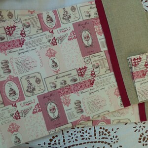 Peut inclure: Un tissu rose et blanc avec un motif répété de macarons, gâteaux et pâtisseries. Le tissu a une bordure rouge et est plié sur un tissu beige.