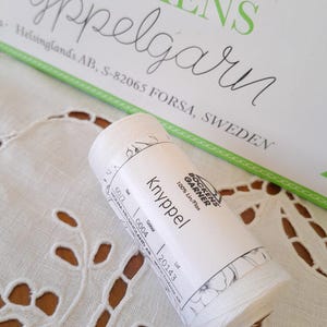 May include: A white spool of Knyppel thread, 100% Lin/Flax, with a label featuring the text "Bockens Garner." The spool is set on a white, embroidered fabric with a floral design. The packaging includes the text "Helsinglands AB, S-82065 FORSA, SWEDEN."