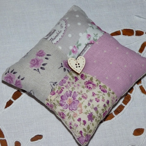 Peut inclure: Un coussin à épingles carré avec un motif patchwork. Il présente des sections grises à pois, des motifs floraux et du tissu lavande uni. Un bouton en forme de cœur en bois est fixé sur le devant. Le mot "Thimble" est imprimé.