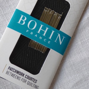 Könnte beinhalten: Eine Packung Bohin France Patchwork-Nadeln. Die weiße Verpackung hat einen türkisfarbenen Streifen mit der Aufschrift "BOHIN FRANCE". Die Packung enthält 10 Nadeln und den Text "PATCHWORK COURTES BETWEENS FOR QUILTING."