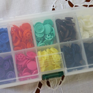 Puede incluir: Una caja de almacenamiento de plástico transparente con 10 compartimentos llenos de botones de plástico de varios colores. Los botones son en tonos de azul, morado, rosa, amarillo, verde, marrón, blanco y negro.