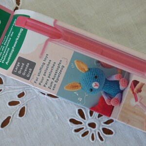 May include: A Clover brand stuffing tool for toys in its original packaging. The tool is a long, pink plastic rod. The packaging features the text 'Stuffing Tool', 'Large', and 'For stuffing toys', along with an illustration of a blue crocheted rabbit.
