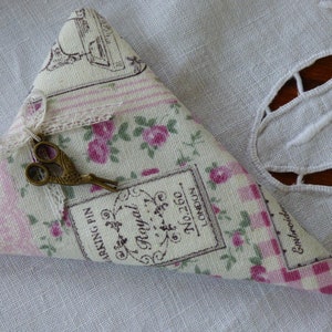 Puede incluir: Un cojín de alfileres triangular de tela con estampado floral y unas pequeñas tijeras de estilo antiguo. La tela tiene un patrón de cuadros rosa y blanco y presenta el texto "Royal" y "No.2260 Londres".