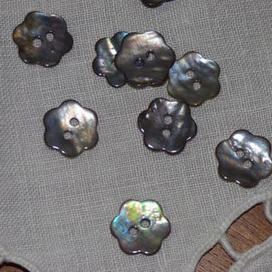 Peut inclure: Plusieurs boutons irisés en forme de fleur, chacun avec deux trous, sont dispersés sur un tissu clair et texturé. Les boutons présentent un éclat nacré, avec des nuances de gris, d'argent et d'autres couleurs.