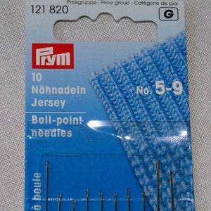Peut inclure: Un paquet d'aiguilles jersey à bout rond de la marque Prym. L'emballage bleu et blanc affiche le texte "10 Nähnadeln Jersey Ball-point needles" et "No. 5-9". Les aiguilles sont visibles en bas du paquet.