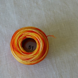 Peut inclure: Une bobine de fil à broder aux couleurs jaune, orange et rouge. Le fil est enroulé serré, avec le numéro "51" visible sur le cœur en carton. Le fond est un tissu beige clair.
