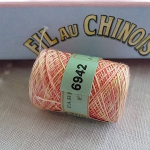 Puede incluir: Una madeja de hilo de bordar rosa y crema con una etiqueta que dice "Fil au Chinois" y "Fabi 6942".