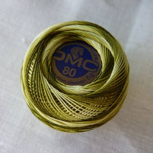 Peut inclure: Une pelote de fil à crochet DMC 80 aux tons verts et jaunes. Le fil est enroulé autour d'une étiquette bleue et dorée indiquant "DMC 80 POUR CROCHET 5g". Le fil est posé sur une surface blanche.