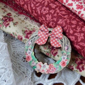 Op de afbeelding: Een decoratieve krans met een roze stippen strik en bloemaccenten rust op een bed van stoffen met patronen. De stoffen omvatten bloemenprints in tinten rood, roze en crème, samen met wit kant.