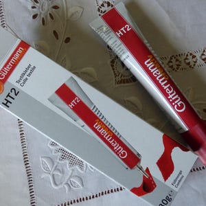 Peut inclure: Un tube de colle textile Gütermann HT2 blanc et rouge accompagné de sa boîte d'emballage en carton assortie. Le produit porte la marque Gütermann, le texte HT2 et l'inscription Textilkleber Colle textile imprimée en noir.