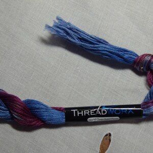 May include: A skein of hand-dyed embroidery floss in shades of blue and purple. The floss is twisted and tied together, and the label reads "THREADWORK Hand Overdyed Floss".
