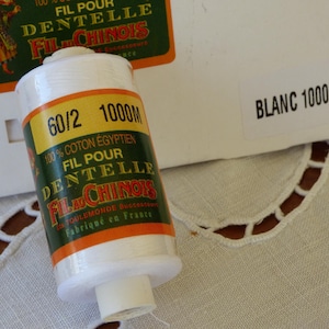 Könnte beinhalten: Eine Spule weißen Fadens mit der Aufschrift "60/2 1000M" in grün und orange. Das Etikett lautet "100% Coton Egyptien, Fil Pour Dentelle, Fil Chinois, Foulemonde Successor, Fabriqué en France". Die Spule liegt auf einer weißen Oberfläche mit einem weißen Etikett, auf dem "Blanc 1000M" steht.