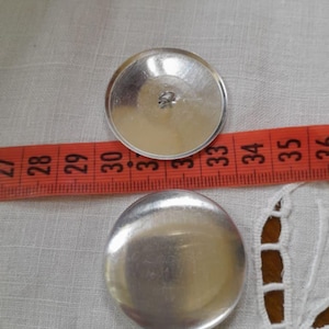 May include: Two silver-colored, round, flat-backed buttons. The buttons are approximately 3.5 cm in diameter.