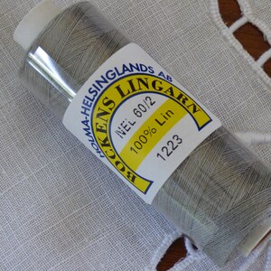 Op de afbeelding: Een spoel grijs linnen garen, met het label "BOCKENS LINGARN" en "100% Lin". Het label is geel en wit. De draad is verpakt in helder plastic. De achtergrond is een witte, getextureerde stof.