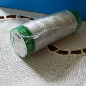 Puede incluir: Una bobina de hilo blanco, envuelta en plástico transparente, con tapas verdes. La bobina tiene una etiqueta con el número "2800". El hilo está sobre una superficie blanca estampada, con un fondo azul.
