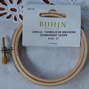 Könnte beinhalten: Ein hölzerner Stickrahmen mit einer Metallschraube und einem Etikett mit der Aufschrift "BOHIN FRANCE EMBROIDERY HOOPS 8 cm - 3". Der Rahmen besteht aus hellem Holz und dient dazu, Stoff beim Sticken straff zu halten.