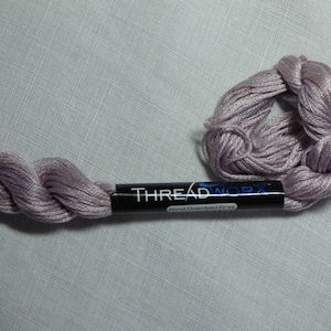 Peut inclure: Une pelote de fil à broder de couleur lavande avec une étiquette noire sur laquelle est inscrit "THREADWORX Hand-Dyed Finish".