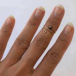 May include: A gold wire ring with a dark pearl accent. The ring is worn on the middle finger of a hand.
