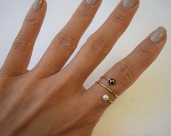 May include: A hand wearing two gold-tone spiral rings, one with a black pearl and the other with a white pearl. The rings are delicate and minimalist, set against a neutral background.
