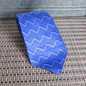 May include: A necktie featuring a repeating geometric pattern of blue and purple squares. Each square contains a series of thin, wavy lines arranged in a grid. The tie has a pointed end and a smooth, slightly reflective fabric texture throughout.