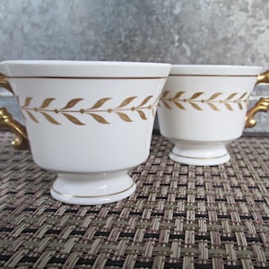 May include: Two white ceramic teacups featuring a gold laurel leaf pattern encircling the upper rim. Each cup has a pedestal base and an ornate, curved gold handle. A thin gold line accents the rim and the base of each cup, providing a cohesive look.
