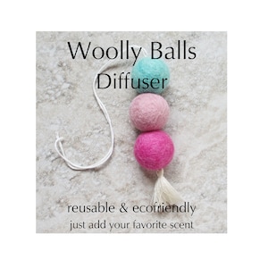 May include: A string of three woolly balls in shades of teal, pink, and magenta, suspended by a white cord. The text "Woolly Balls Diffuser" is above the balls, with "reusable & ecofriendly just add your favorite scent" below.