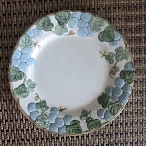 May include: A round ceramic plate with a white center and a scalloped rim featuring a raised, embossed pattern of light blue grapes and green leaves. The design encircles the border of the plate, showcasing detailed textures and natural colors.