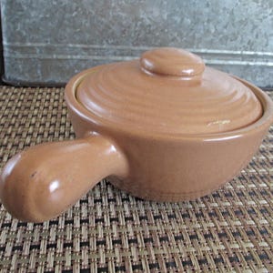 May include: A light brown ceramic individual casserole dish with a matching lid and a single side handle. The round pot features a smooth, solid-colored finish and a series of concentric circular ridges on the top of the lid for a textured look.