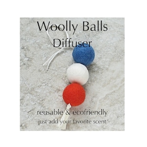 May include: A Woolly Balls Diffuser featuring three felt balls in red, white, and blue, strung together with a white cord. The text reads "reusable & ecofriendly just add your favorite scent."