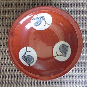 May include: A round, terracotta-colored ceramic bowl featuring a smooth, glossy finish. The interior of the bowl is decorated with three distinct white circular patches, each containing a hand-painted blue and brown snail illustration.