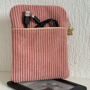 Può includere: Una custodia per e-reader in velluto a coste rosa antico con cerniera dorata e un ciondolo dorato. La custodia contiene un cavo USB nero e un cavo di ricarica nero. Un e-reader è visibile sotto.