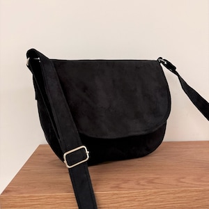 Può includere: Una borsa a tracolla in pelle scamosciata nera con una lunga tracolla regolabile e una fibbia argentata. La borsa ha una chiusura a lembo e una forma arrotondata.