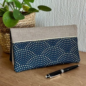 May include: A rectangular clutch bag with a navy blue star pattern and a tan linen top. A black pen with silver accents rests on a wooden surface. A small potted plant in a woven basket is in the background.
