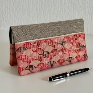 Peut inclure: Une pochette rectangulaire avec un rabat. La pochette présente un motif de demi-cercles roses et rouges superposés. Le rabat supérieur est de couleur beige uni. Un stylo noir avec des accents argentés repose sur la surface.