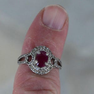 May include: A silver ring featuring a large oval ruby gemstone, encircled by smaller clear stones. The ring has a decorative design on the band and around the central ruby. The ring is on a finger.