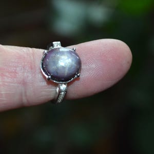 May include: A silver ring with a large, oval, purplish-grey gemstone. The gemstone has a star-like effect. The ring's band is decorated with small, clear stones. The ring is held up by a finger.